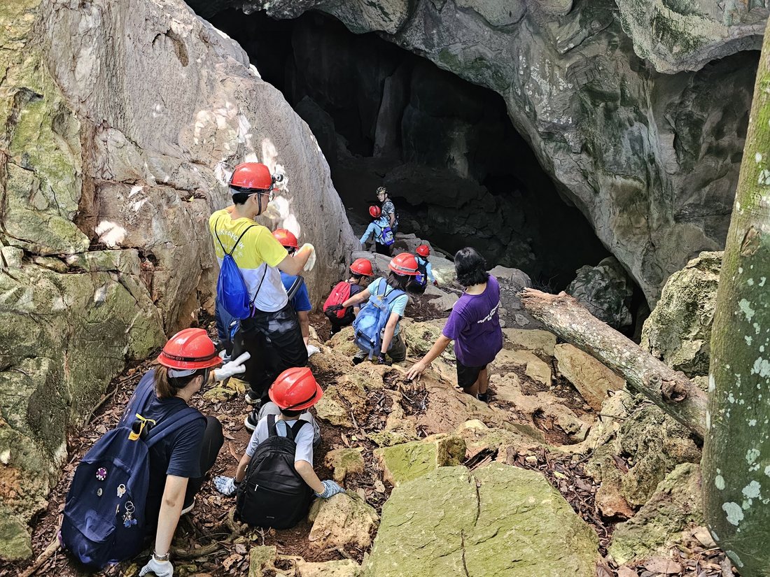 Students in red helmets climbing rocks towards a cave entrance